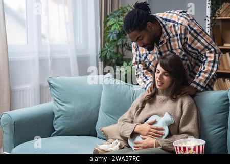 Coppia di razza mista uomo afro-americano e donna bianca, maschio massaggia la sua fidanzata o moglie durante il periodo mestruale mentre si siede a casa. Amore, suppo Foto Stock