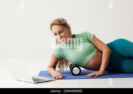 ragazza che tiene in mano un rullo massaggiante seduto sul pavimento e vicino a un computer portatile, rullo massaggiante per il fitness e lo yoga, allenamenti online a casa Foto Stock