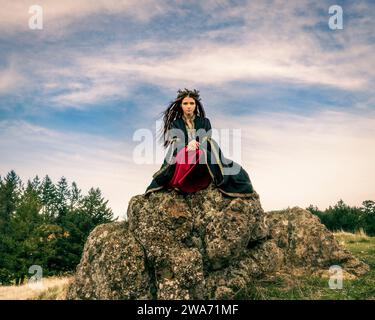 Giovane donna Strega pagana in Inverno Pine Floral Crown Mountain Peak Nuvolose Sdraiate Rocks pomeriggio Solstizio Day Magico Foto Stock