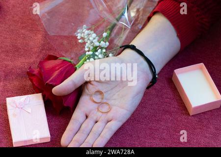 mano che regala anelli di fidanzamento con una confezione regalo aperta e una rosa rossa sullo sfondo Foto Stock