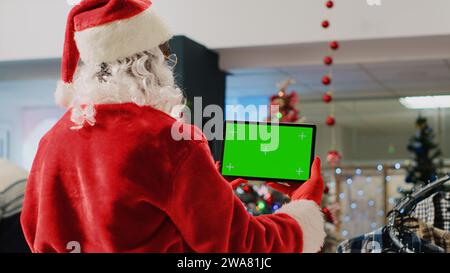 Dipendente di un centro commerciale ornato di Natale che impersona Babbo Natale con un tablet mockup, che si prende una pausa dal lavoro per guardare video sullo schermo dei tasti cromatici mentre i clienti girano e si avvicinano Foto Stock