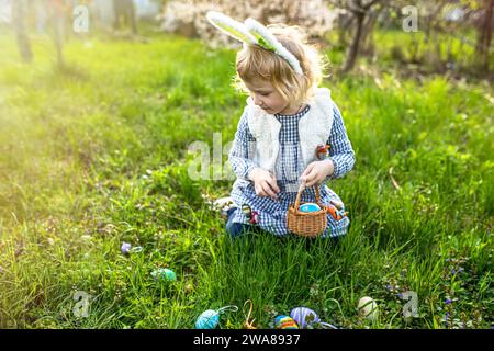 La bambina indossa orecchie da coniglio e raccoglie uova di Pasqua colorate durante la caccia all'uovo di Pasqua in giardino Foto Stock