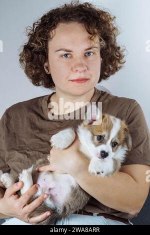 Ritratto di giovane attraente donna dai capelli castani con capelli ondulati in T-shirt in cotone naturale, che tiene in mano un divertente e soffice cucciolo di vellshkorgi, Foto Stock