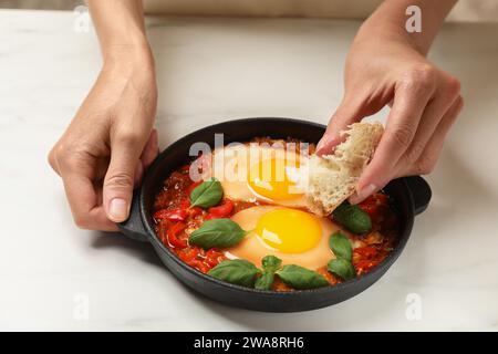 Donna che immerge un pezzo di pane nel delizioso Shakshuka al tavolo bianco, primo piano Foto Stock