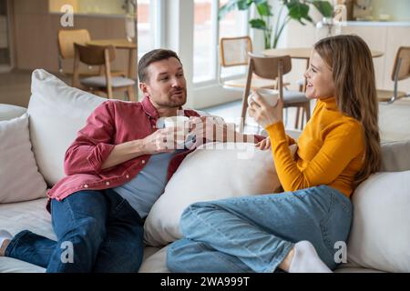 Coppia allegra e rilassata seduta sul divano, chiacchierando, discutendo, ridendo, gustando il tè a casa Foto Stock