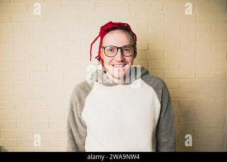 giovane e attraente hipster con dreadlocks rossi in occhiali e maglione bianco sullo sfondo di una parete in mattoni. Foto Stock