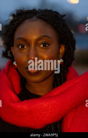 Questo ritratto cattura una giovane donna nera in posa avvolta da una brillante sciarpa rossa. L'immagine trasuda calore e raffinatezza, evidenziata dal delicato effetto bokeh delle luci della città sullo sfondo. Lo sguardo del soggetto è diretto verso la fotocamera, con un sorriso sottile e sicuro. Il suo trucco naturale accentua i suoi tratti e gli orecchini donano un tocco di eleganza. L'illuminazione accentua la tonalità della pelle e la consistenza dei suoi capelli, creando un contrasto armonioso con le fredde tonalità blu del crepuscolo sullo sfondo.il crepuscolo risplende di eleganza. Foto di alta qualità Foto Stock