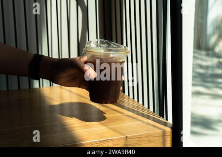 Mano che tiene il caffè di ghiaccio nero in un bicchiere da bere su uno sfondo di legno con un fascio di sole dalla finestra. Drink rinfrescante estivo. Caffè nero in Foto Stock