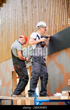 Carpentieri che costruiscono casa incorniciata in legno. Due operai che si accerchiano la facciata della casa con pannelli in cemento, fissandoli con cacciaviti. Concetto di moderna costruzione ecologica. Foto Stock