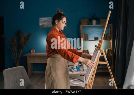 Artista femminile creativa latina in piedi che lavora su un nuovo dipinto nel suo laboratorio domestico. Felice giovane donna che dipinge su tela nel suo studio d'arte. Foto Stock