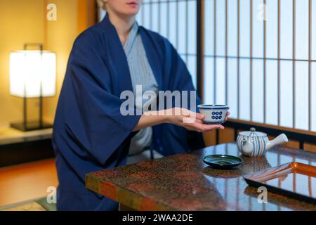 Giovane donna caucasica in abiti giapponesi seduta al tavolo nella sala ryokan, beve un tè Foto Stock