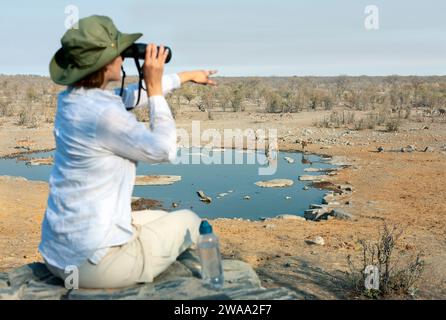 Vista posteriore di una giovane donna bionda in safari seduto sulla roccia guardando attraverso il binocolo - concetto di viaggio Foto Stock