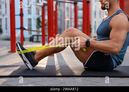 Uomo muscoloso che fa esercizio fisico con fascia elastica di resistenza sul tappetino in campo sportivo, primo piano Foto Stock