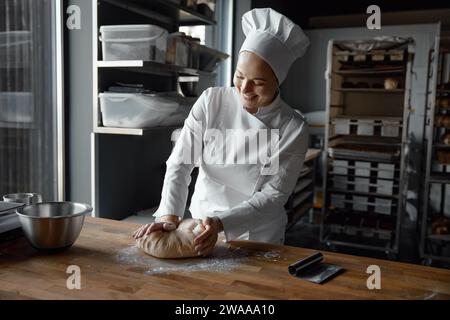 Una giovane donna fornaio felice e attraente che indossa l'uniforme per impastare l'impasto in cucina Foto Stock