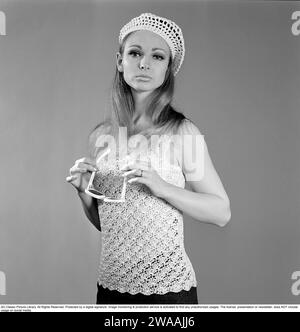 Negli anni '1960 Una giovane donna in uno studio di fotografi ha fotografato indossando un maglione senza maniche a maglia, lavorato a maglia in un modo arioso. Il tappo è lavorato a maglia nello stesso maniero. Sta indossando gli occhiali da sole. 1967. Foto Stock