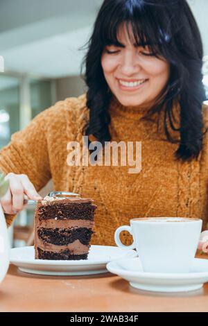 donna caucasica sorridente sfocata seduta tagliando una fetta di torta al cioccolato, concentrati sulla torta con una tazza di caffè sul tavolo. Foto Stock