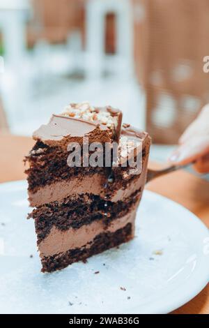 immagine verticale della mano di una donna caucasica irriconoscibile che taglia con una fetta di torta al cioccolato servita su un piatto di porcellana bianca su un tavolo di legno, pa Foto Stock