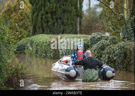 Worcester, 3 gennaio 2024 - Jack, di 7 anni e mezzo, viene trasportato in barca alla sua casa nel borgo inglese di Severn Stoke, completamente tagliato dalle inondazioni dopo che il fiume Severn ha fatto scoppiare le sue rive a causa delle continue piogge della tempesta Henk. Il pub Rose and Crown potrebbe essere visto pompare acqua mentre combatteva per rimanere asciutto con le loro difese fatte in casa contro le inondazioni. - Credito: Stop Press Media/Alamy Live News Foto Stock