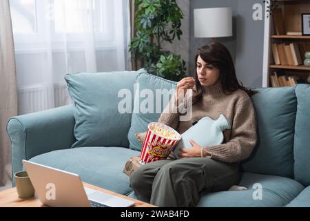 Giovane donna con mal di stomaco o mestruazioni seduto sul divano a casa a mangiare popcorn e a guardare film online sul computer portatile. Doloroso problema mestruale, fe Foto Stock