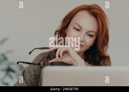 Donna professionista dai capelli rossi che tiene gli occhiali, guarda lo schermo del notebook con un sorriso contemplativo Foto Stock