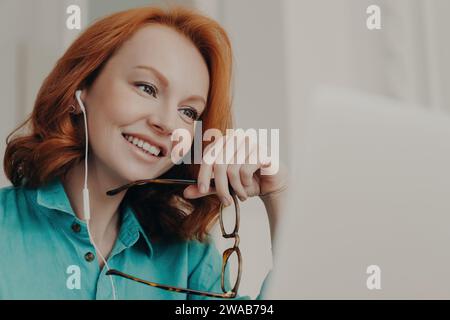 Allegra donna dai capelli rossi con auricolari che reggono occhiali e che partecipa a una videochiamata su un computer portatile Foto Stock