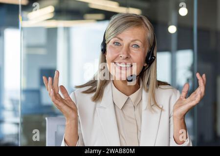 Ritratto ravvicinato di una donna d'affari anziana seduta in un auricolare in ufficio di fronte alla fotocamera, che parla al telefono, gestisce con le mani. Foto Stock