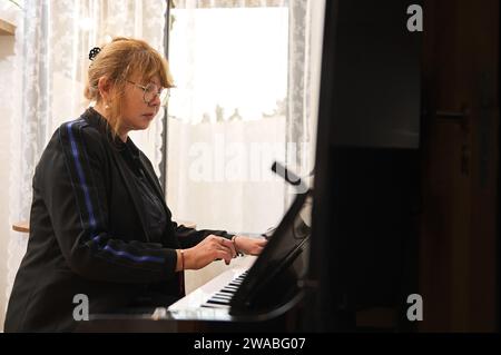 Autentica donna asiatica matura sicura di sé in un abbigliamento casual nero, che suona il pianoforte e canta, insegna lezioni di pianoforte a casa Foto Stock