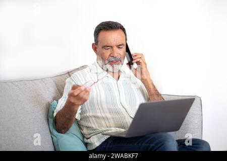 Uomo anziano che parla al telefono per risolvere i problemi con il notebook in interni Foto Stock
