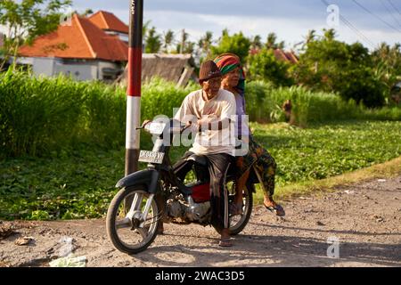 Una pittoresca coppia di anziani che cavalca una motocicletta su una strada in un villaggio di Bali. Bali, Indonesia - 12.10.2022 Foto Stock