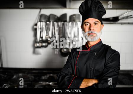 Cuoco maturo in cappotto nero con braccia piegate nella sua cucina Foto Stock