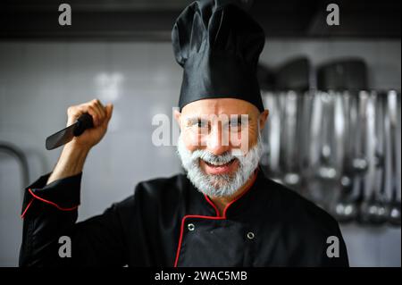 Chef arrabbiato con barba bianca in un cappotto nero che tiene un coltello che ti minaccia Foto Stock