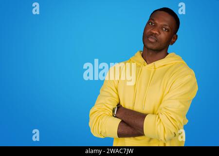 Un uomo africano sicuro di sé sta con le braccia incrociate e indossa una felpa gialla con cappuccio Foto Stock