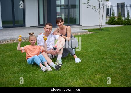 Madre, padre e figlia felici seduti sull'erba e guardando la macchina fotografica in cortile Foto Stock