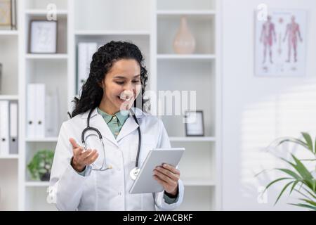 Il medico all'interno dello studio medico è in piedi con un tablet computer in mano, un operatore clinico in veste medica bianca consiglierà il paziente da remoto utilizzando il video per videochiamata, con gioia e amicizia. Foto Stock