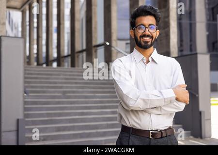 Ritratto di un uomo d'affari dall'esterno dell'edificio degli uffici, un giovane uomo di successo sorride e guarda la macchina fotografica con le mani incrociate, un impiegato in abiti da lavoro. Foto Stock