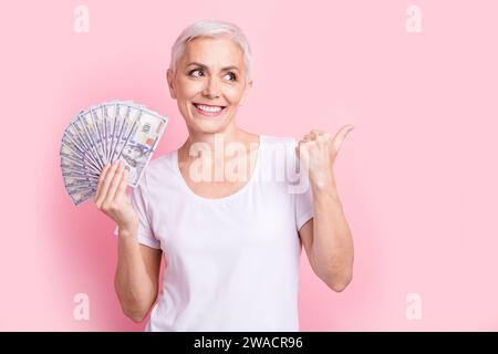 Foto di attraente donna pensionato tenere soldi fan point guardare spazio vuoto vestiti eleganti vestiti bianchi isolati su sfondo di colore rosa Foto Stock