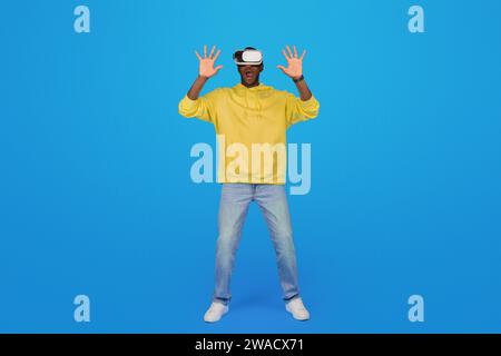 Un uomo nero con una felpa gialla con cappuccio rimane stupito con le mani alzate e indossa occhiali per realtà virtuale Foto Stock