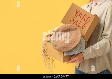 Donna che tiene una scatola di oggetti indesiderati e un cartello di cartone con testo IN VENDITA su sfondo giallo Foto Stock