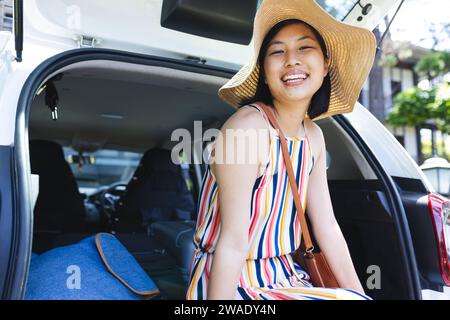 Ritratto di donna asiatica felice seduta in macchina e con cappello da sole in giardino a casa. Vita domestica, estate e stile di vita, inalterato. Foto Stock