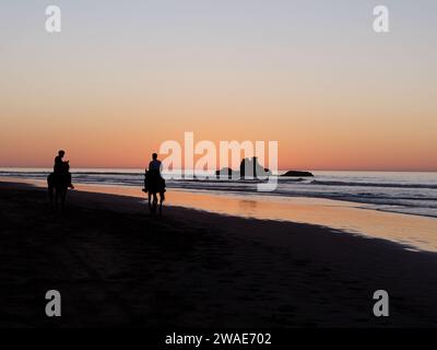 I cavalieri da spiaggia al tramonto in silhouette con un affioramento roccioso mentre la luce arancione si riflette nell'acqua. Essaouira, Marocco, 3 gennaio 2024 Foto Stock