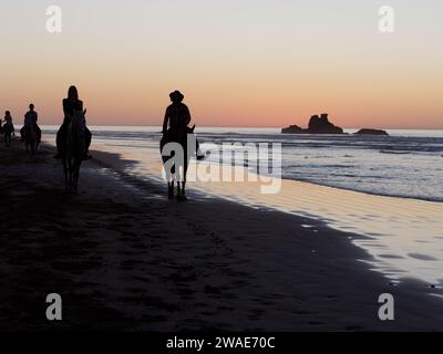 I cavalieri da spiaggia al tramonto in silhouette con un affioramento roccioso mentre la luce arancione si riflette nell'acqua. Essaouira, Marocco, 3 gennaio 2024 Foto Stock