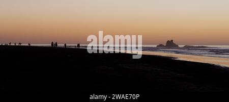 I cavalieri da spiaggia al tramonto in silhouette con un affioramento roccioso mentre la luce arancione si riflette nell'acqua. Essaouira, Marocco, 3 gennaio 2024 Foto Stock