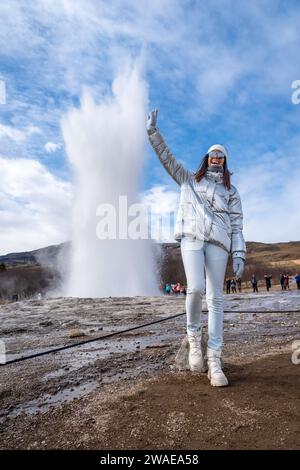 Una giovane donna vestita di bianco in piedi di fronte a un geysen geyse circondato da una vegetazione lussureggiante Foto Stock