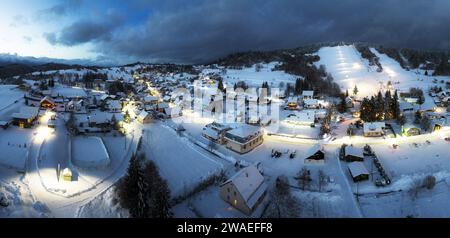 Vista della catena montuosa innevata e del villaggio con case illuminate situate in fondo alla collina di Slovkia al tramonto Foto Stock