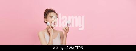 Giovane donna attraente che applica un cuscino sul viso con polveriera su sfondo rosa Foto Stock