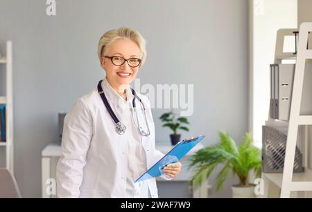 Ritratto di felice bello anziano medico femminile con clipboard in piedi in ufficio medico Foto Stock