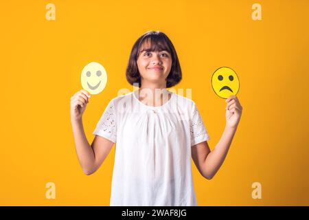 La ragazza tiene in mano emoticon tristi e felici. Concetto di salute mentale, psicologia ed emozioni dei bambini Foto Stock