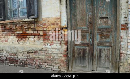 frammento della parete esterna di un'antica casa con una porta in legno malandata e una finestra ricchissima, con un angolo in mattoni distrutto come una vecchia struttura Foto Stock