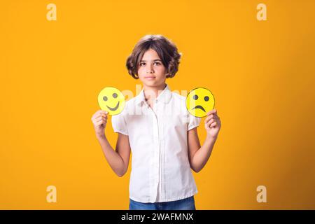 La ragazza tiene in mano emoticon tristi e felici. Concetto di salute mentale, psicologia ed emozioni dei bambini Foto Stock