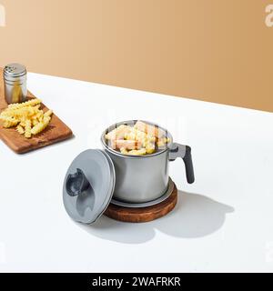 Vassoio dell'olio in acciaio inox, con patatine fritte e salsiccia in cima e su un vassoio di legno, su sfondo bianco e beige. Foto Stock